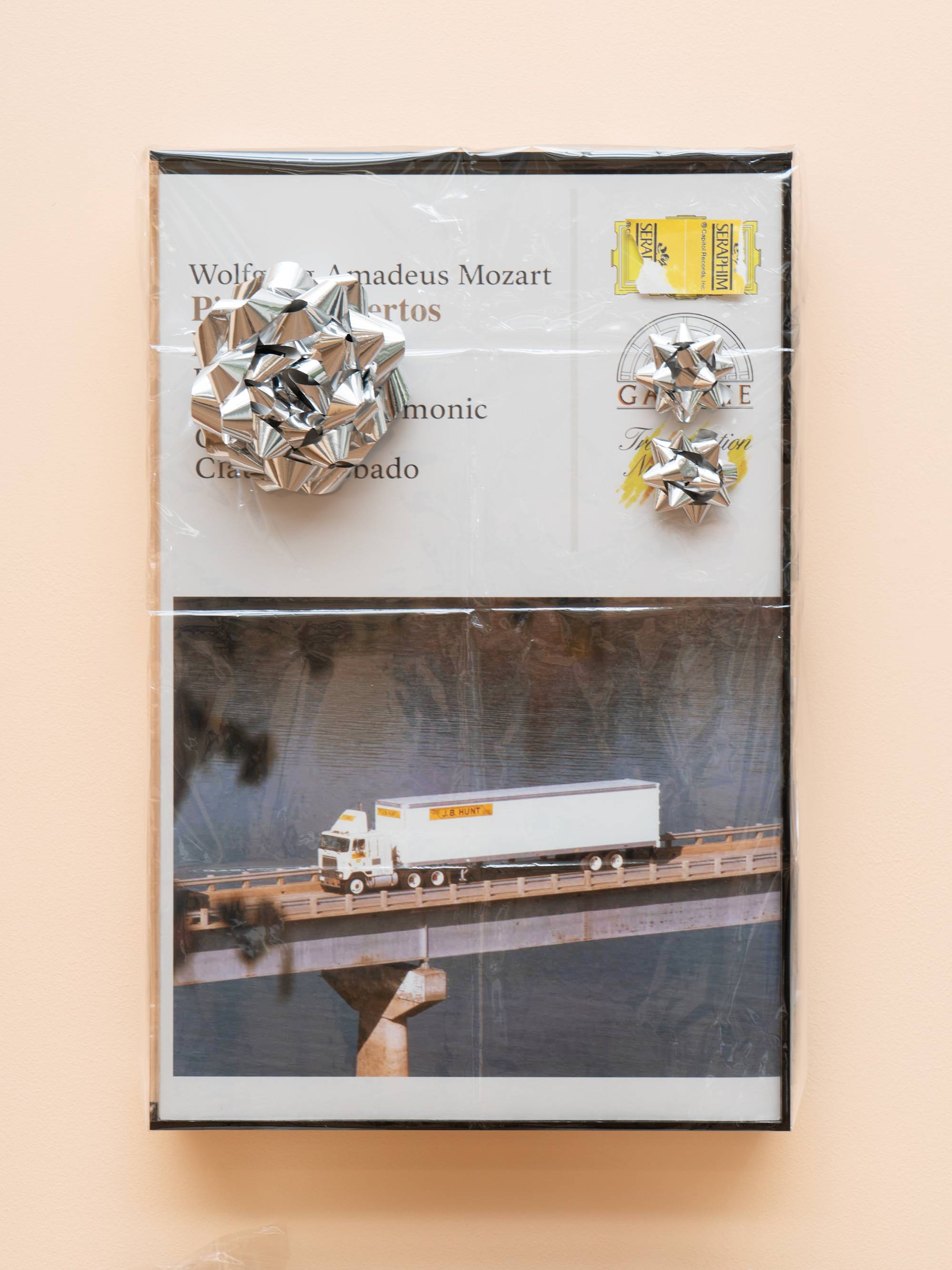 Detail of a wood veneered shelf hangs on a peach colored wall. 2 tan, plaid, circular gift boxes sit on the shelf with tan socks in plastic bags draped over them. A small drawer juts out of the front of the shelf and is neatly filled with trimmed business cards. A large, fake audio cassette case featuring a truck driving over a bridge hangs above the shelf, wrapped in cellophane and decorated with three silver gift bows.
