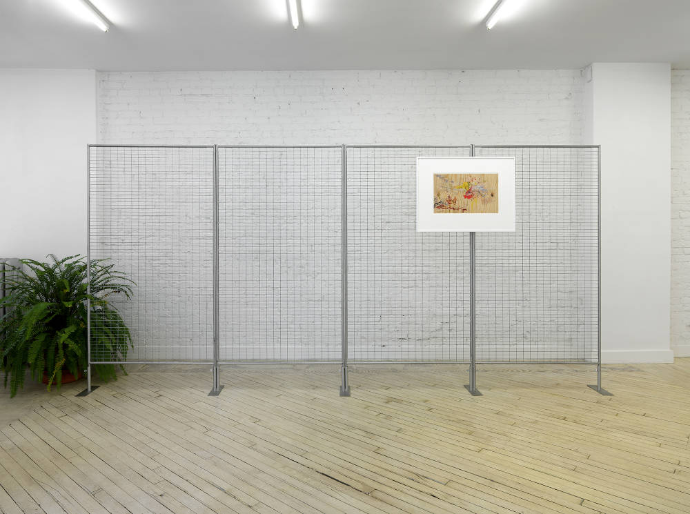 In a gallery space, A large multi-part chain link fence is freestanding in front of a brick wall painted white. To the right side of the chain-link fence hans a framed drawing depicting a series of abstract marks in numerous colors on a tan ground. The frame is white.To the left of the fence is a planted fern. 