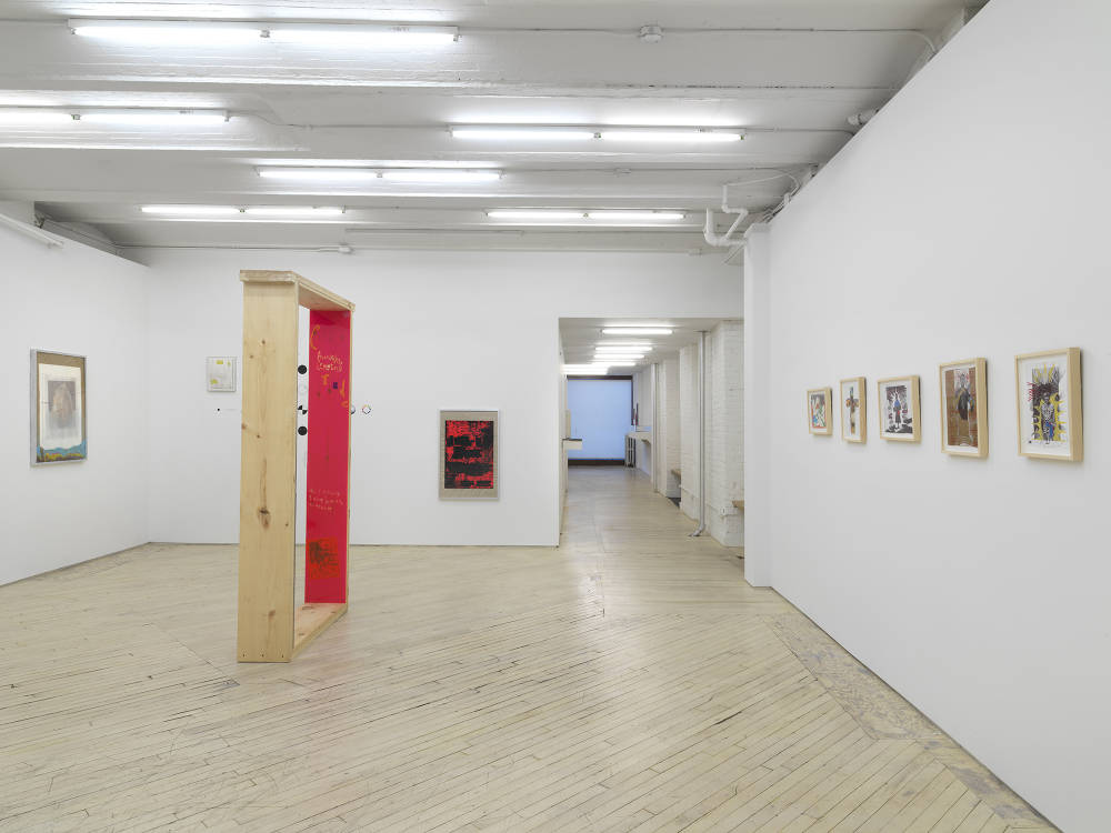In a gallery space, a row of small framed drawings. In the center a large wooden sculpture with a bright pink side. In the background is an installation of framed photographs at various heights. 
