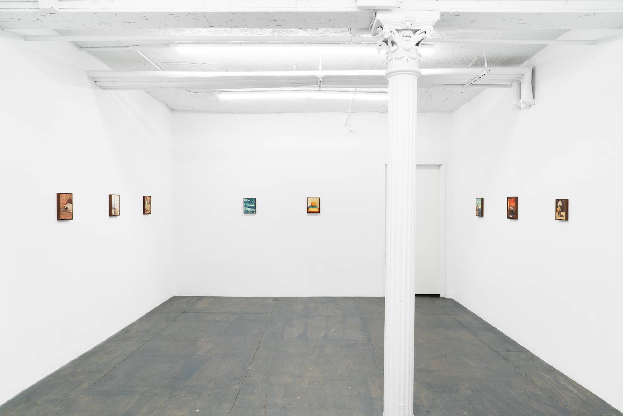 Wide shot of a white-walled art gallery with several small paintings displayed on the walls. A large white pillar stands in the center of the room, and the floor is grey painted wood.