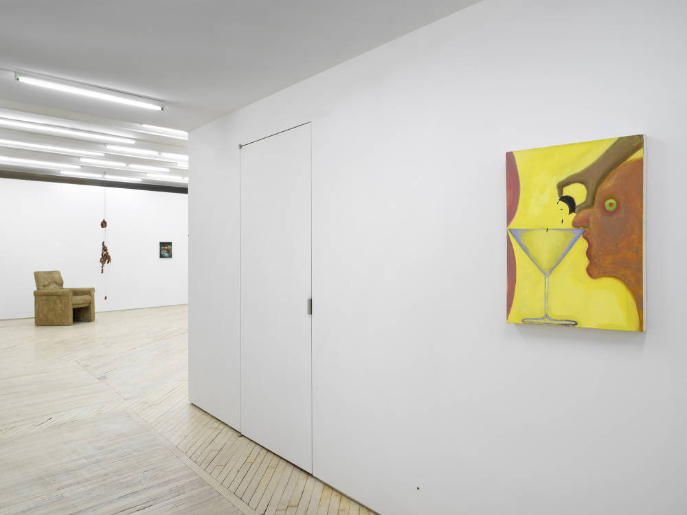 In a gallery space, a bright yellow and orange painting depicts the profile of a face leaning against the brim of a martini glass. In the background is a large sculpture of a brown chair with a sculpture consisting of sausages links beside it suspended from the ceiling. There is a small, darkish painting to the right. 