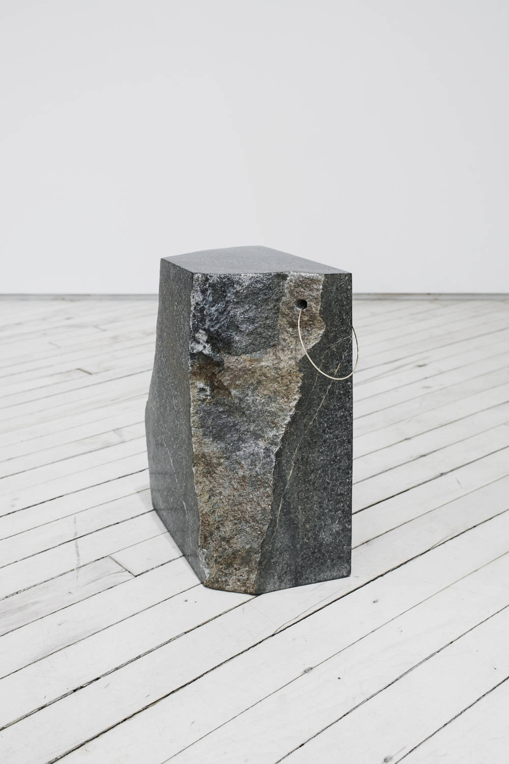 A large stone with natural edges sits on a wooden gallery floor, the stone has a hole drilled through the top right corner where a large hoop earring pierces the stone.