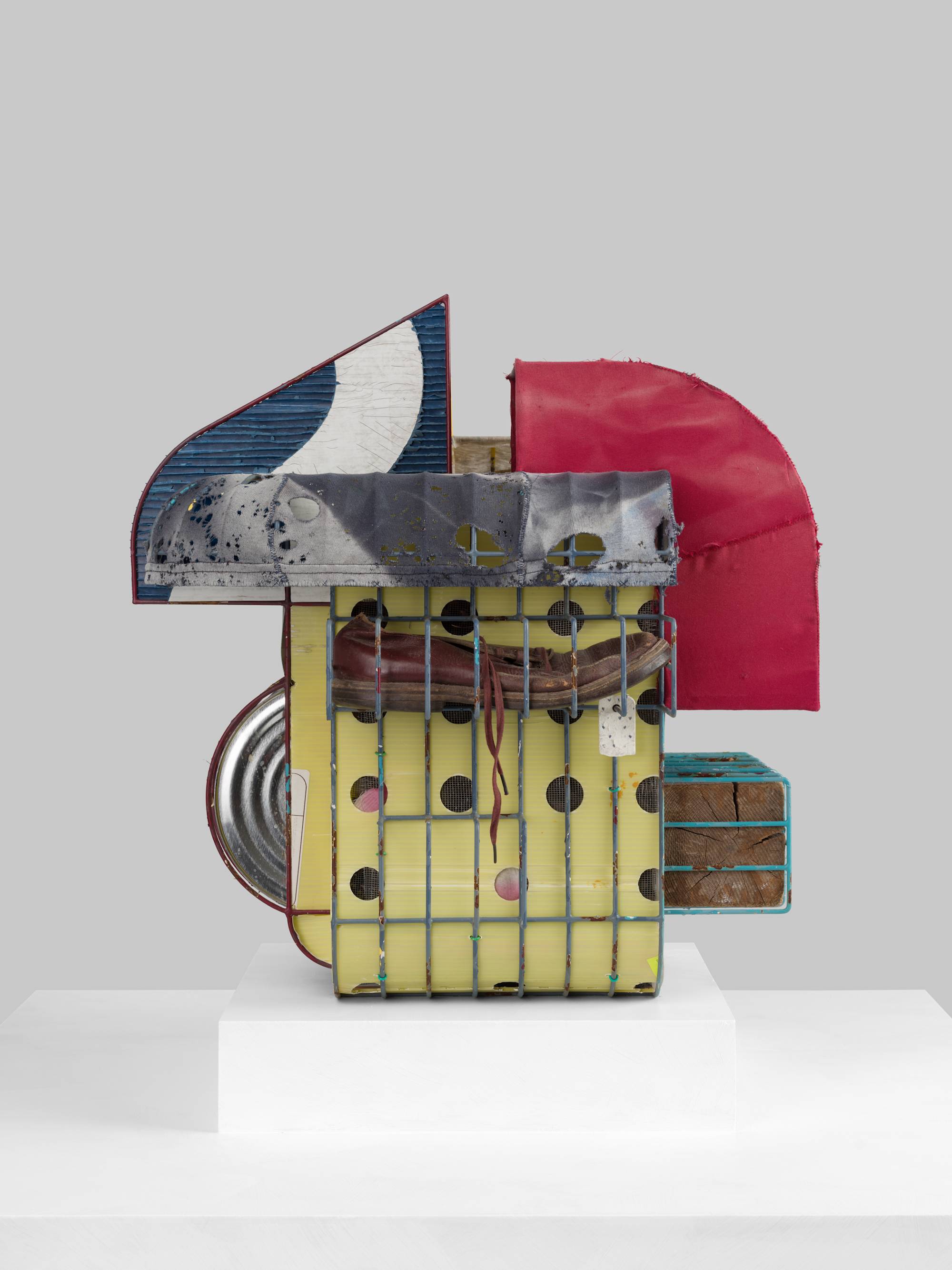 A cage-like colorful steal wire sculpture, wrapped with several tattered fabrics to resemble various awnings, and embellished with an old shoe, a twig, printed coroplast signage, an old blue paint can, a lock, a paper tag, and a wooden block sits on a white pedestal against a white wall.