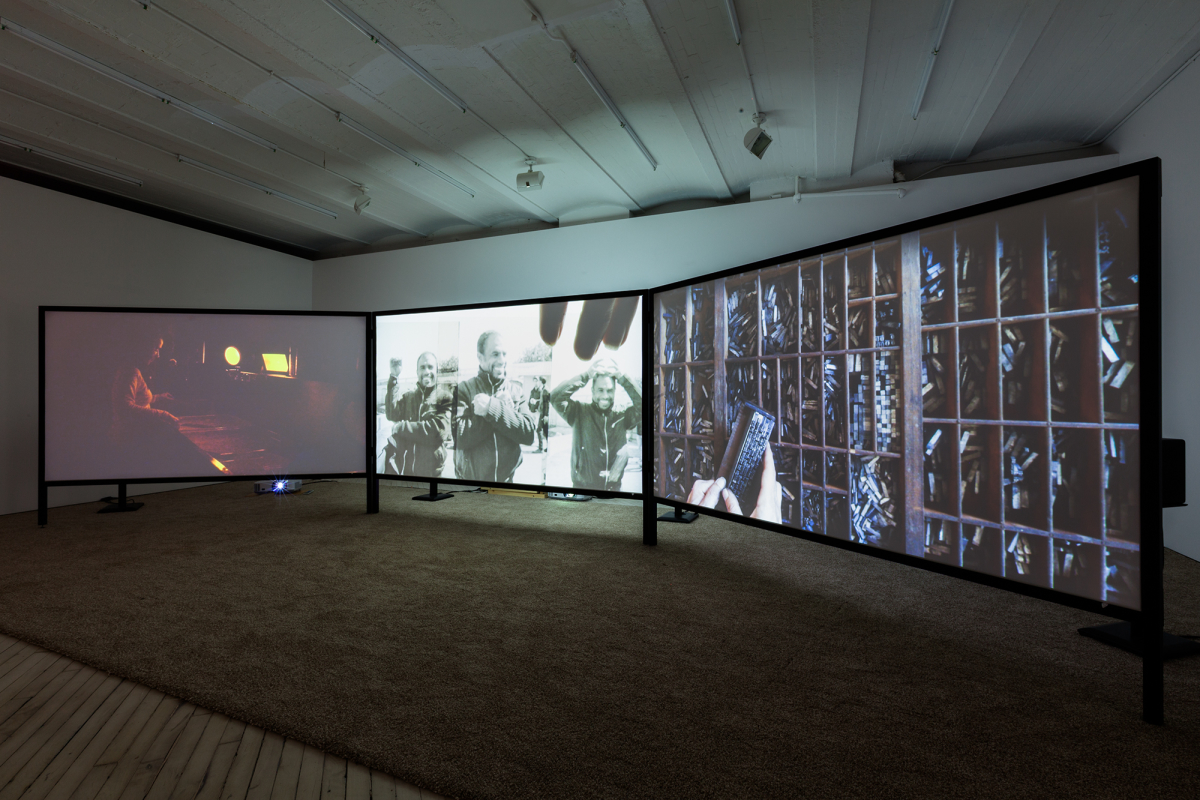 Image of the darkened gallery - a three screen installation of a video. At left the artist is seen in a darkroom, developing a photograph, the center shows a black and white image of a man making hand gestures and at right a close up of a wooden case for the lead block letters for typesetting print.
