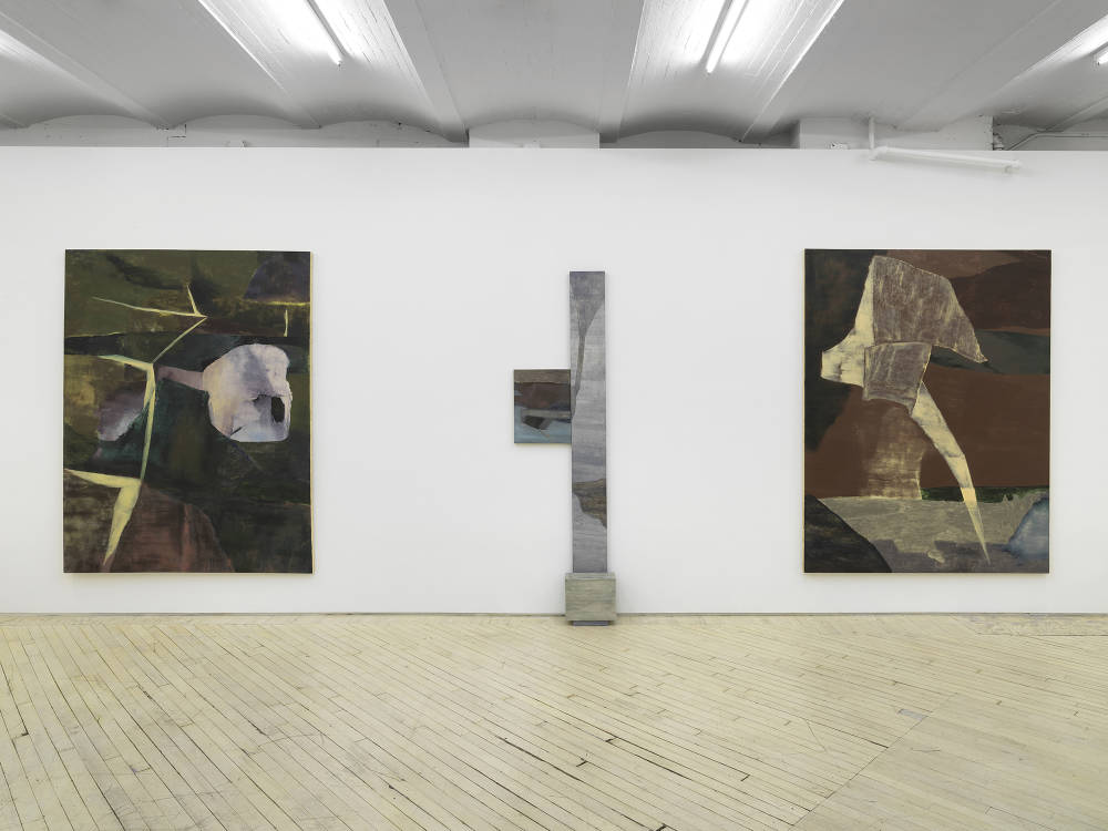 Two large abstract paintings installed in a gallery with a vertical leaning painting and sculpture in the center. Each are rendered in muted palette of browns and greens, predominantly. 