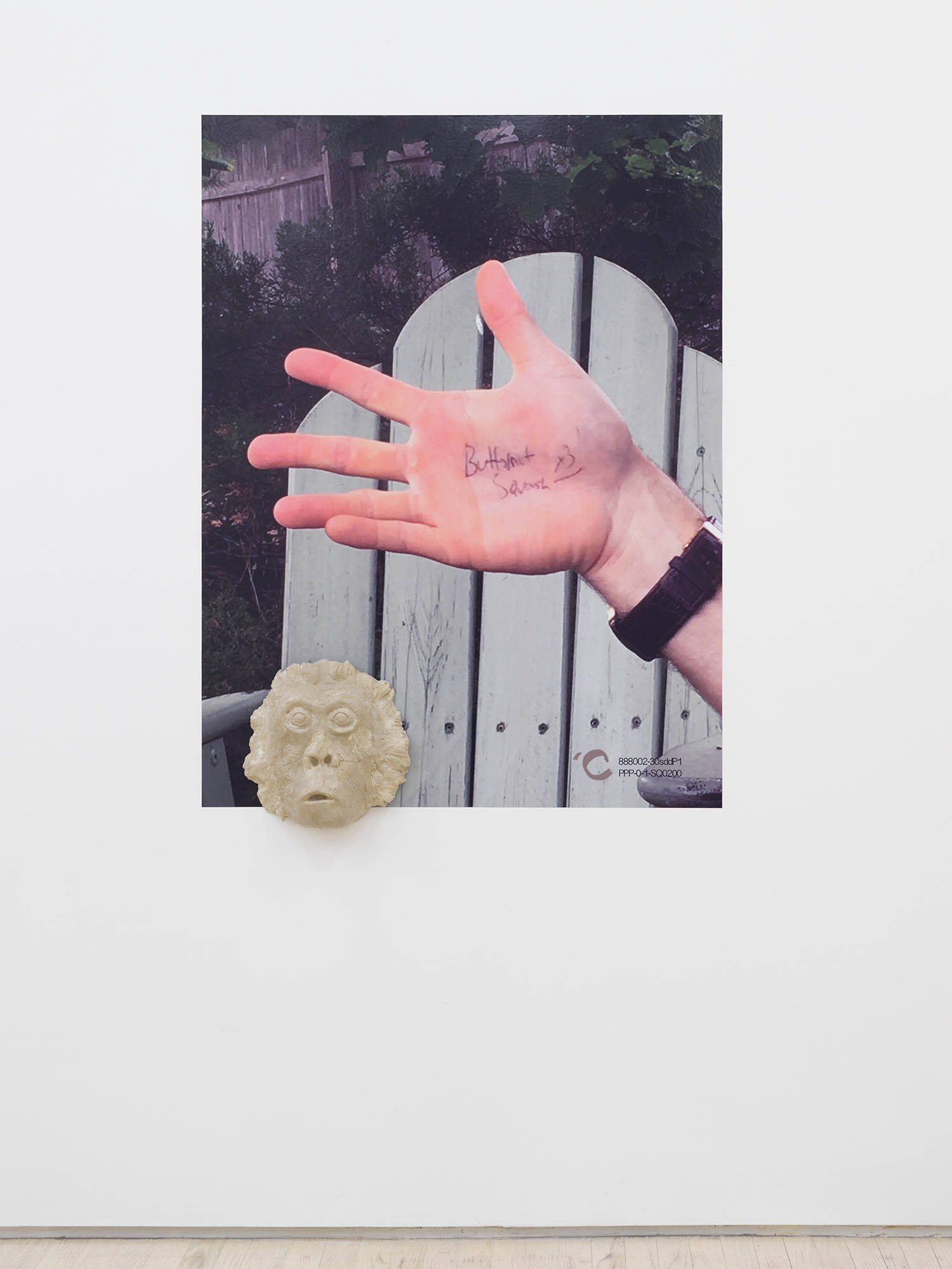 Image of a photographic artwork of a hand with writing on the palm in front of a lawn chair. On the bottom left of the artwork is a clay mask installed.