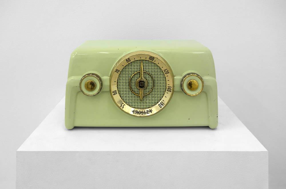 On top of a white pedestal is vintage green radio with golden accents placed in front of a white wall. 