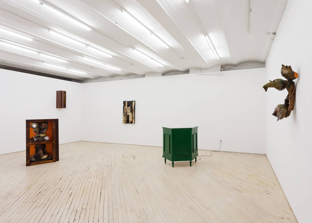 Installation view of an exhibition by Brandon Ndife. In a gallery there are two sculptures sitting on the floor appearing like pieces of furniture or cabinetry. Hanging on the wall are three other sculptures each appearing like a sconce, a piece of floor, and a kitchen cabinet.