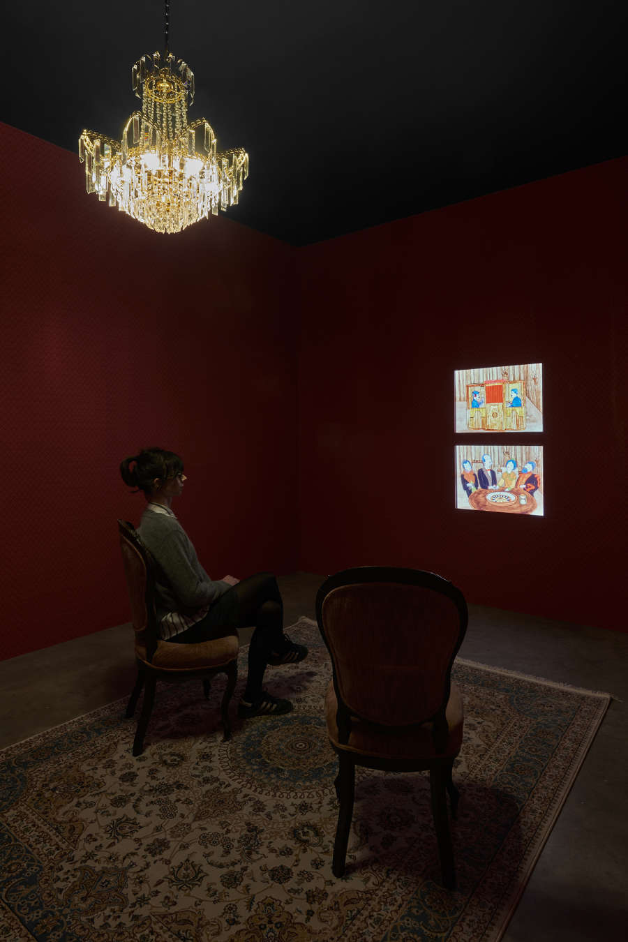 Installation view of a red room with a chandelier and a person sitting in a chair in front of two small projections on the wall in front of them.
