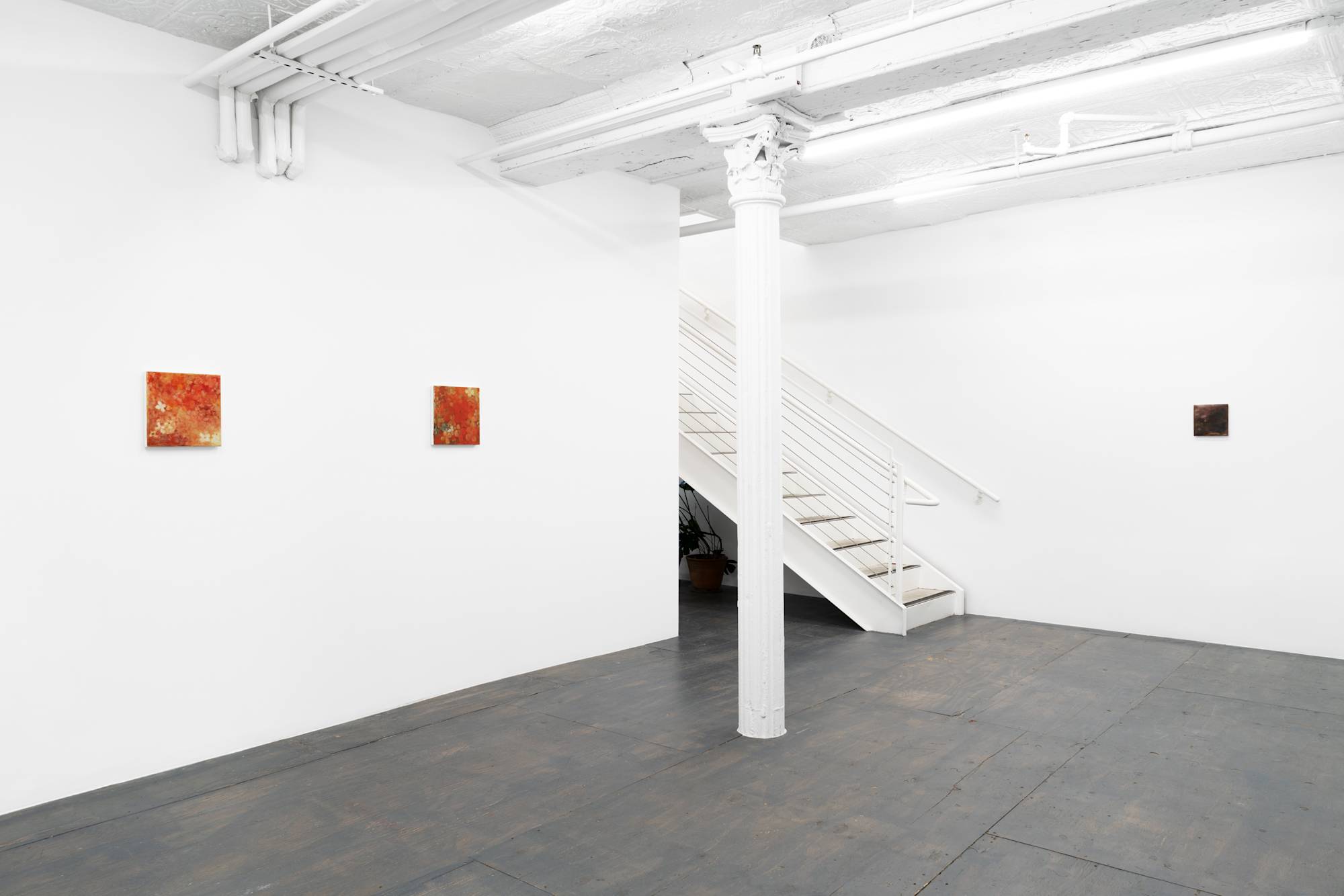 Three small abstract paintings of flowers featuring various colors spaced evenly in the corner of a brightly lit gallery with grey wood floor. There is a white column in the center of the gallery and stairs leading to another floor, out of view.