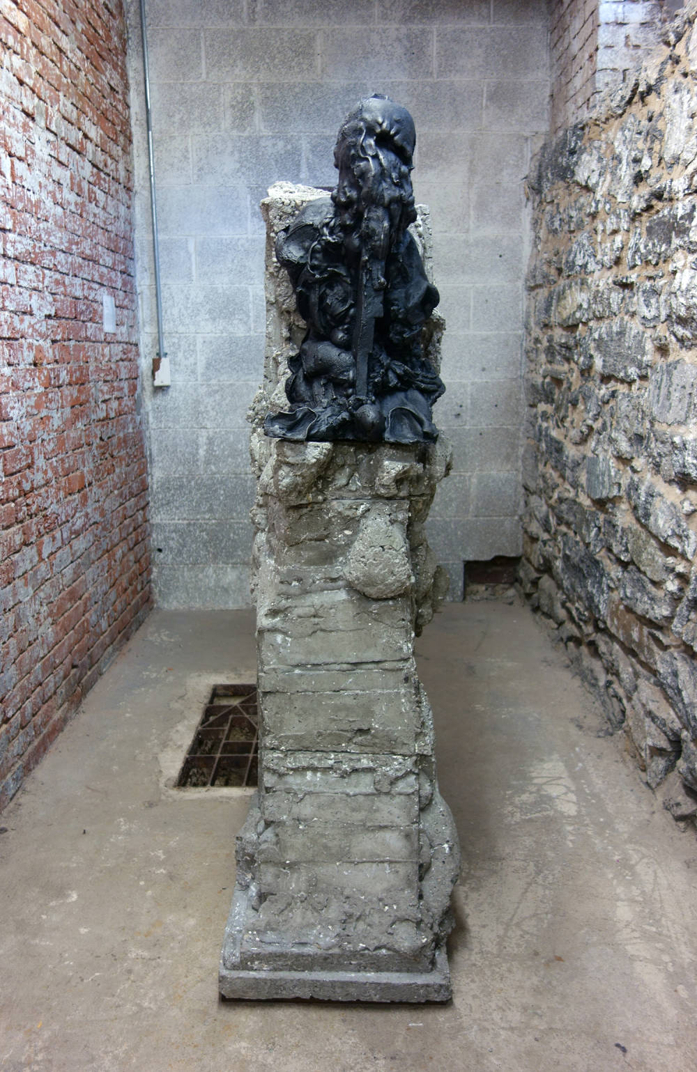 Installation view of metal and concrete sculpture, depicting a cast iron sculpture of a head and torso melting and/or decaying atop a jagged concrete plinth.