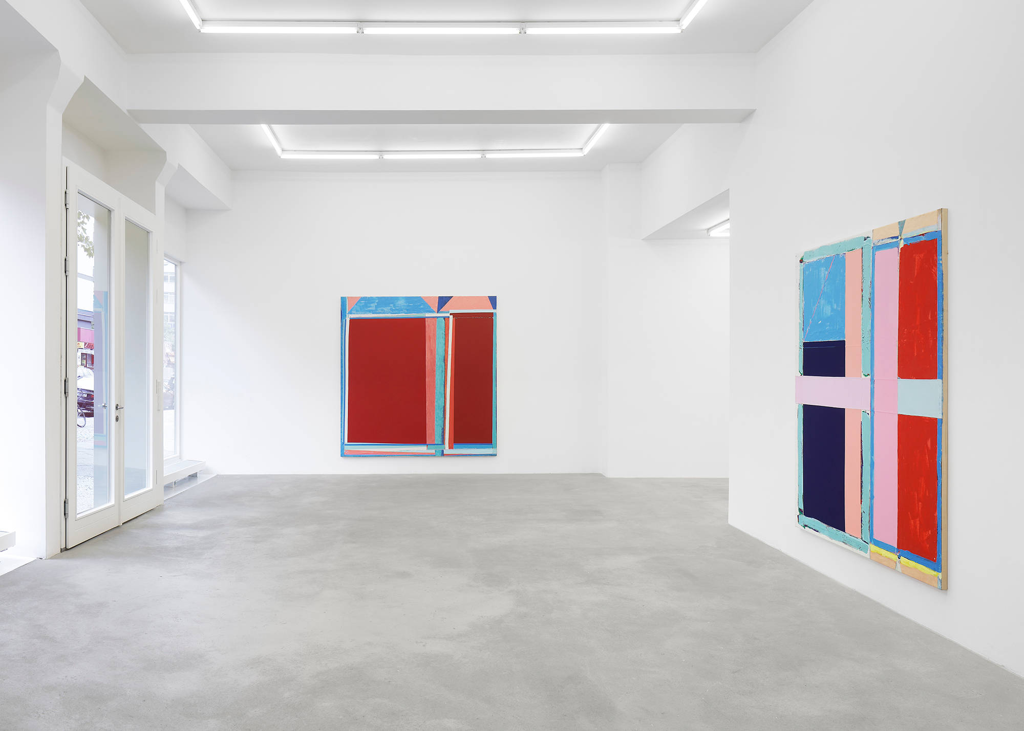 In a gallery space, two large abstract paintings hung on separate walls. The paintings are formally dominated by large rectangular blocks of color. The most striking colors in both paintings are a range of reds, pinks, and blues. The paint is very thick and the canvas is rough.
