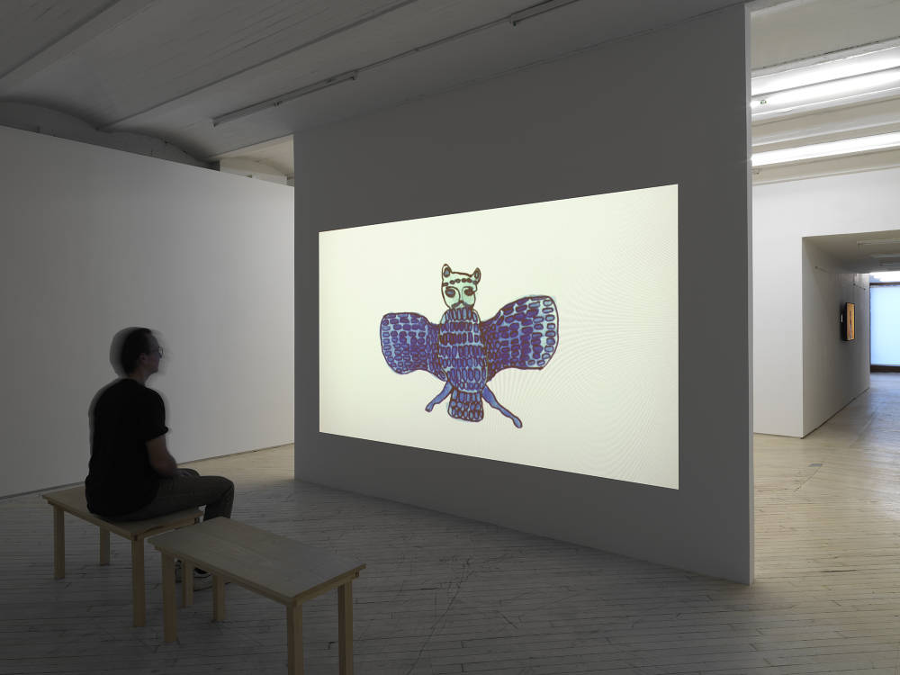 A film is projected in a gallery, the screen bright white with an ink drawing of a blue owl like bird, a person sitting on a bench watching the film.