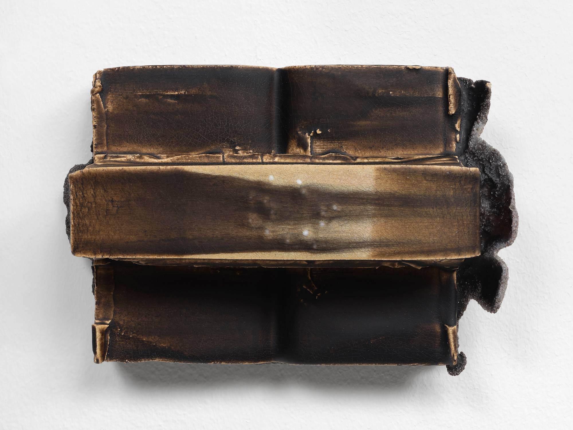 A small, brown, ceramic sculpture featuring two vertical cuboids with a long horizontal band stretching the full width embedded with a circle of white beads.