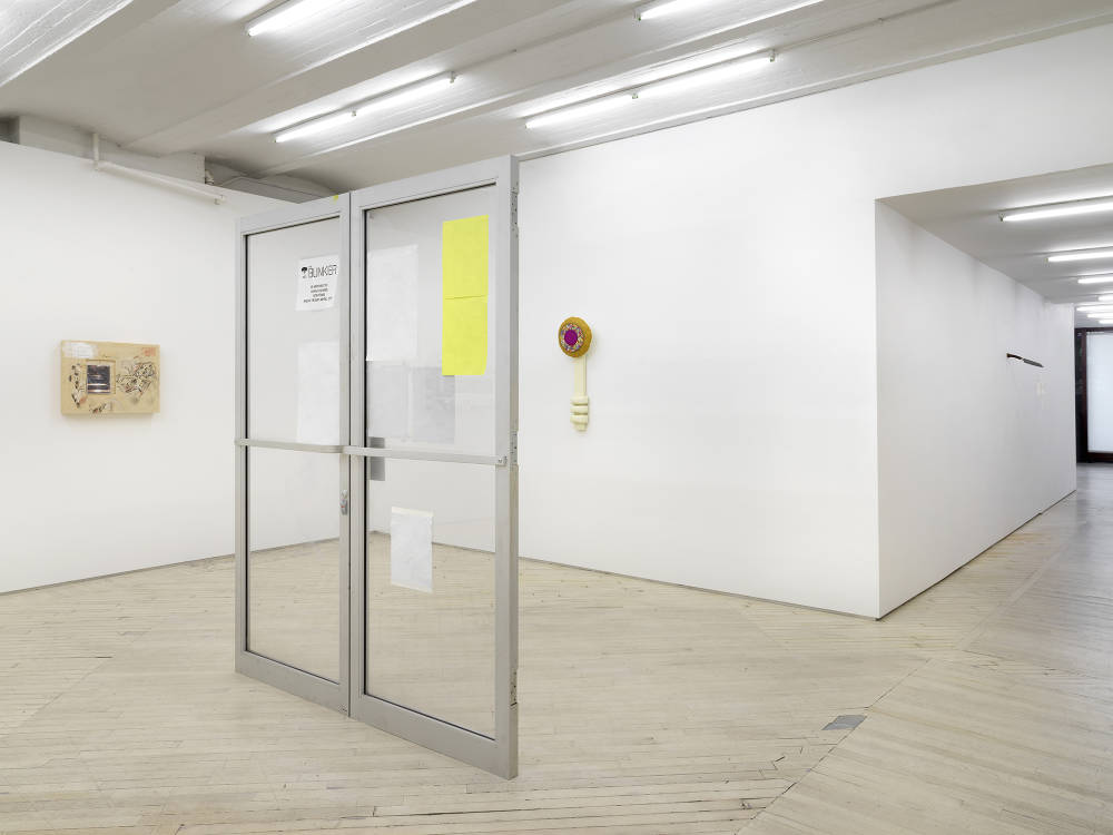 In a gallery space, a large freestanding metal and glass doorway sculpture. Attached to the glass are multiple poster advertisements.In the background are two wall sculptures. To the right is a long hallway with a large window at the end. 