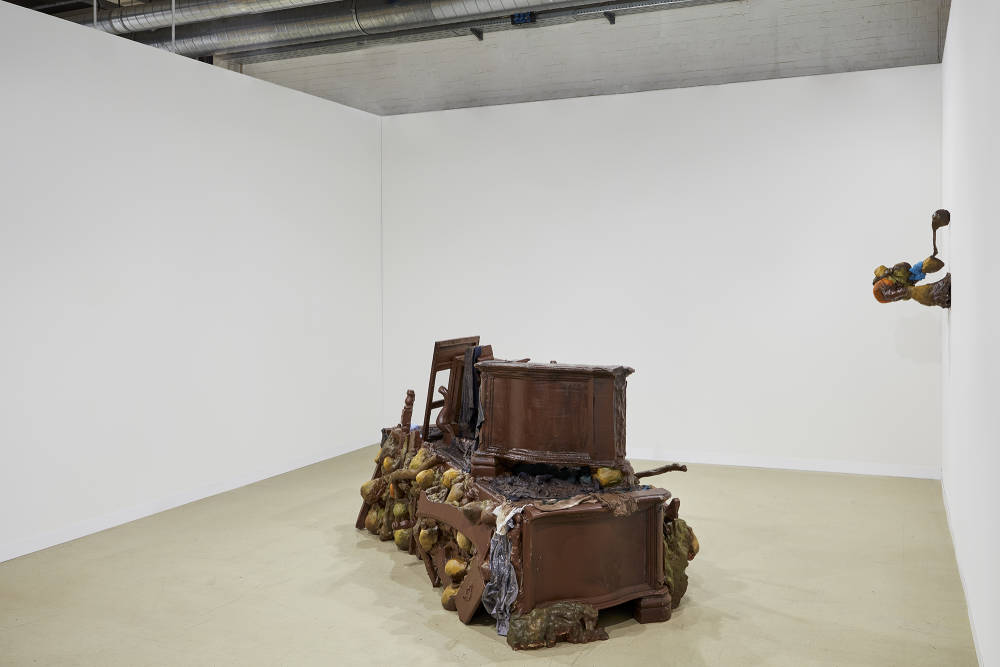 A large piece of brown furniture sits in the middle of a room with white walls. A smaller sculpture hangs to the right of the larger installation on the wall.