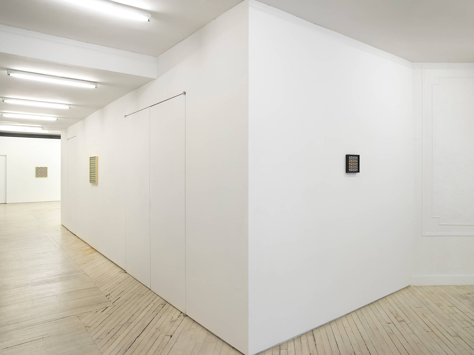 In a gallery space, a small painting hangs on a wall in a black frame depicting a repeating geometric pattern. In a hallway is a larger painting also a repeating geometric pattern with a yellow frame. There is a similarly sized painting in the background in a larger gallery space.