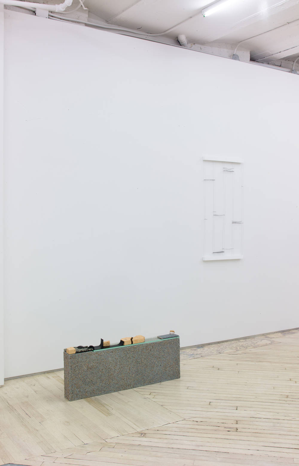 In a gallery space are two objects. The object in the foreground is a small, rectangular, architectural structure, resting on the floor. To the right is an abstract, wooden object hung on a white wall. 