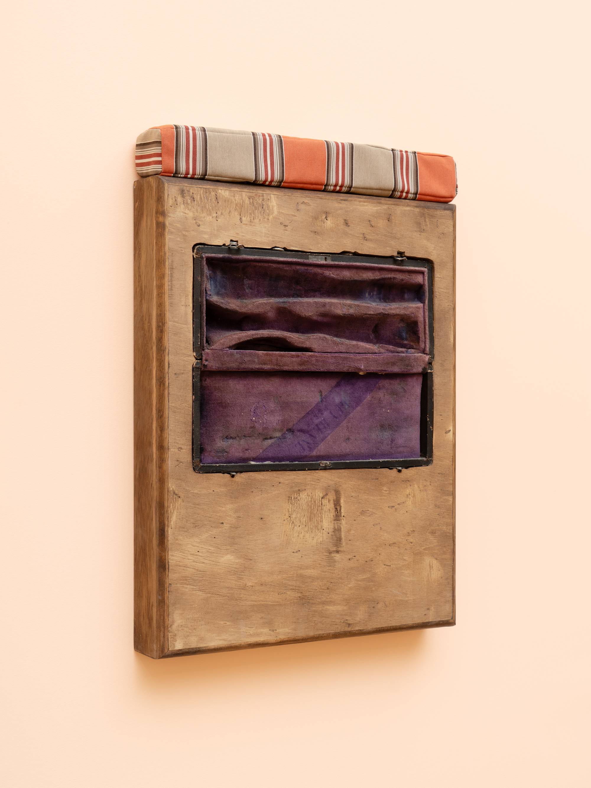 Quarter view of a wooden box sculpture with inset clarinet case with purple velvet lining, topped with a orange and beige striped cushion, hanging on a peach colored wall.