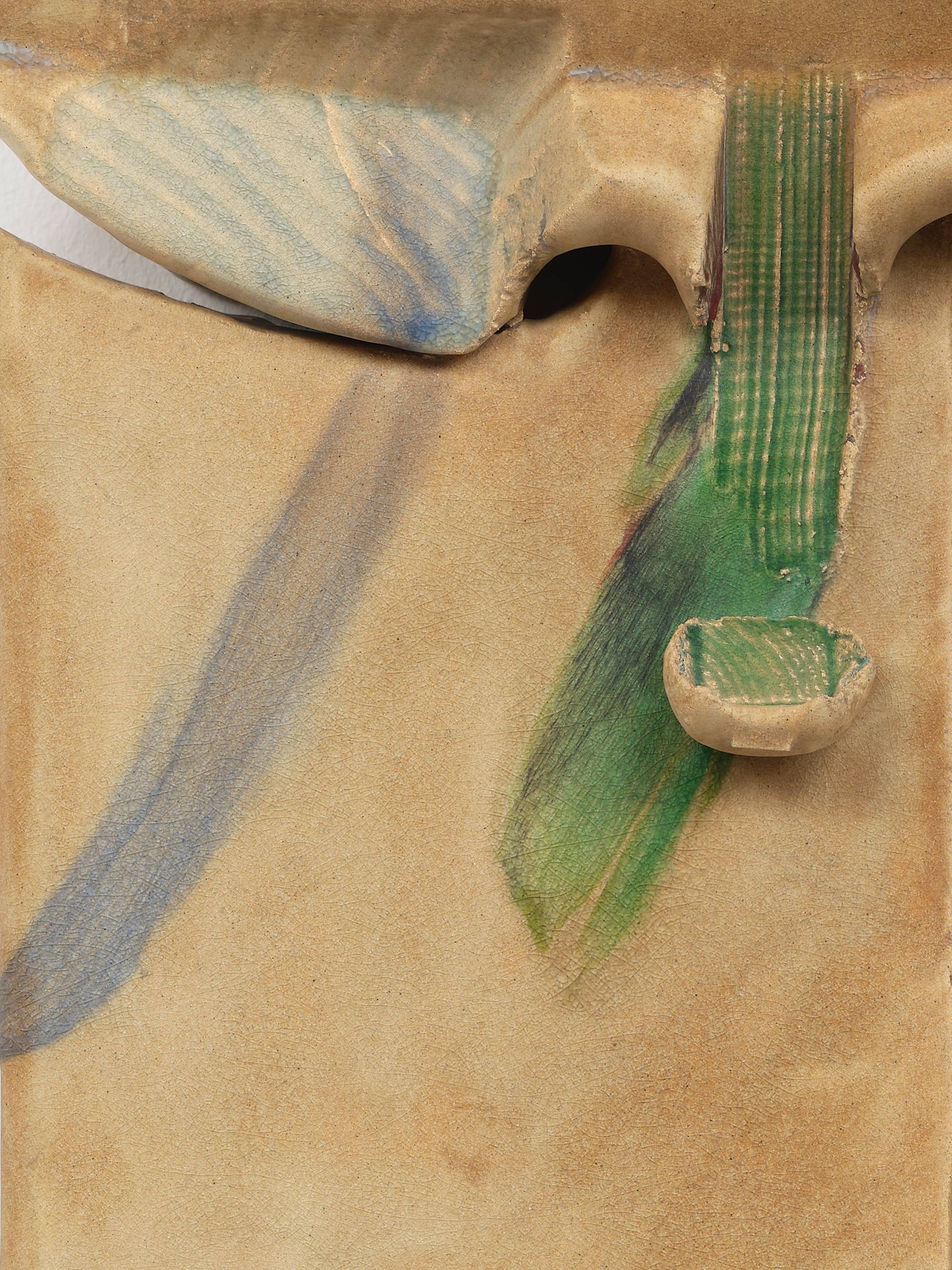Detail of two ceramic sculptures hang on a wall mirroring each other with a thin channel dividing them. The clay has been pushed and stamped to create appendages near the upper edges that resemble small wings glazed faintly blue. Towards the center of the composition, both pieces have been gouged in a downward motion through the shoulder of the wings resulting in narrow vertical troughs that end in a small lifted flaps. A green glaze runs through the troughs and flaps. Each sculpture is tipped with a small peninsula still coated with sand from the kiln they were fired in.