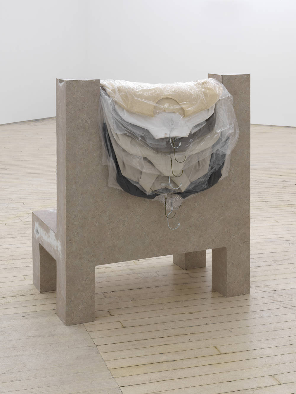 In a gallery space, a sculpture rests in the center of the room. The object resembles an architectural structure with several pale colored shirts in dry cleaning bags draped over the top. 