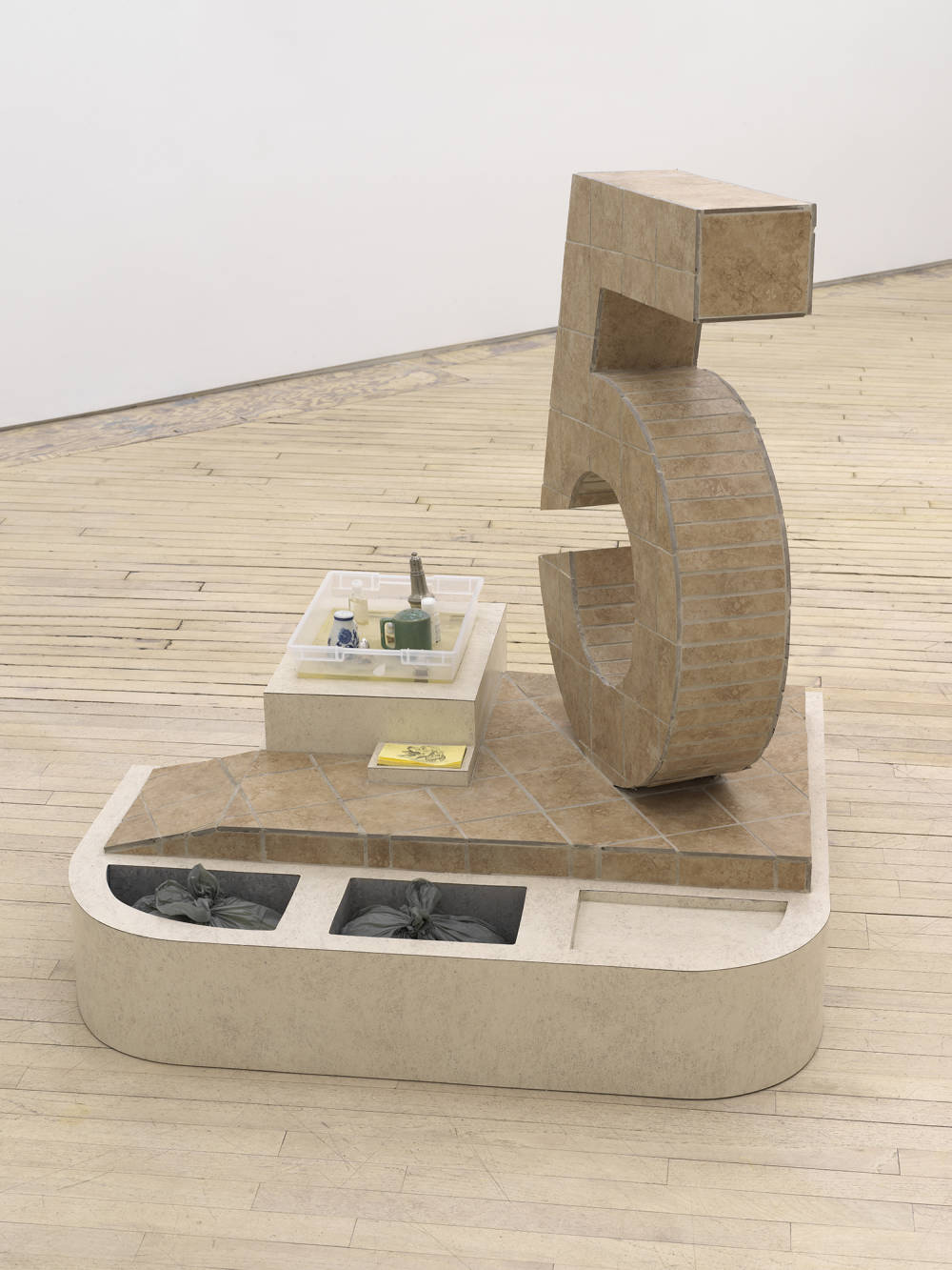 In a gallery space, a large abstract number 5 sculpture constructed out of flooring titles. The base of the sculpture contains two plastic bags and several salt and pepper shakers. There is a stack of post-it notes.