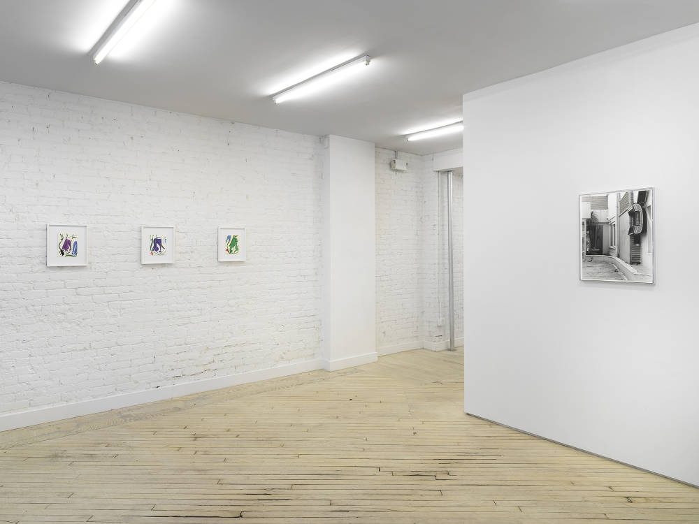 Image of the front room of Bureau gallery containing a white wall with a black and white framed photograph of a telephone booth on an empty street. On the left there are three colorful framed drawings by Patricia Treib