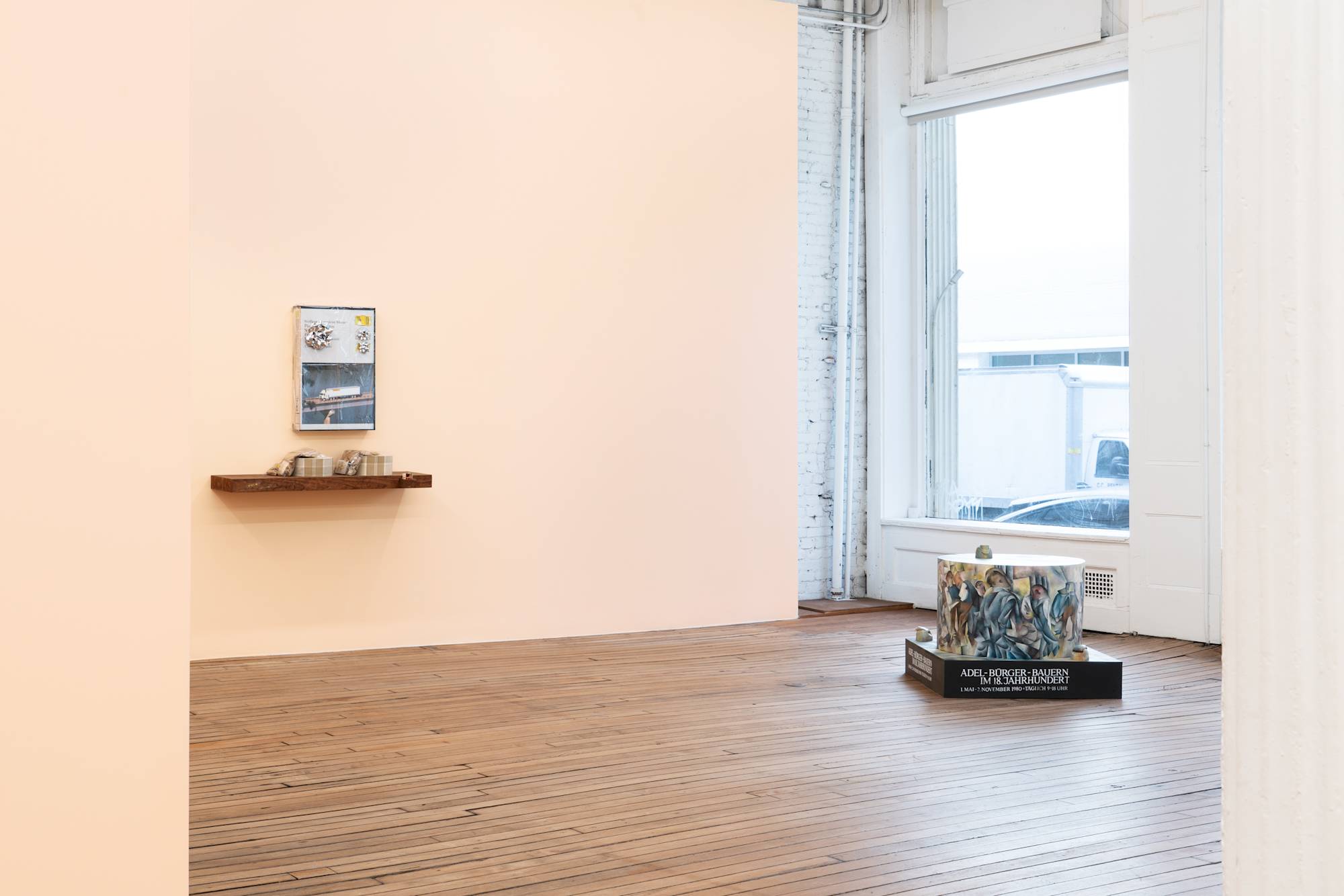 A freestanding sculpture with a round center sits in a gallery space in front of a window. The sculpture is painted in a Cubist style. To the left, a wood veneered shelf with an oversized fake cassette case hang on a peach colored wall