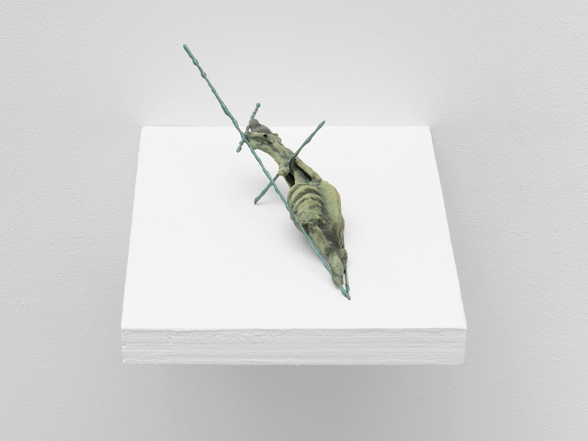 A small, handmade sculpture by Matt Hoyt sits on a white ply wood shelf. It resembles a light green snail on a dark green cross.