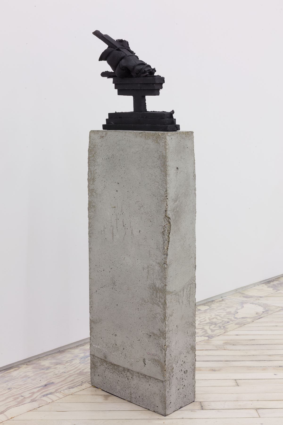 Installation view of cast iron and concrete sculpture resting on the floor, depicting a tiered geometric form on top of a tall concrete plinth.