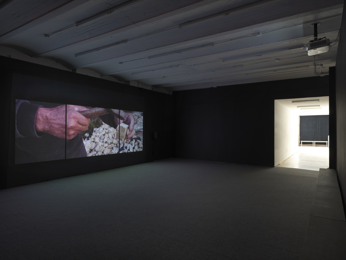 View of Gallery space with Video Installation projected onto dark wall, which exits into a lit hallway. Projected is a still of a hands shaping a piece of limestone with a small hammer. 