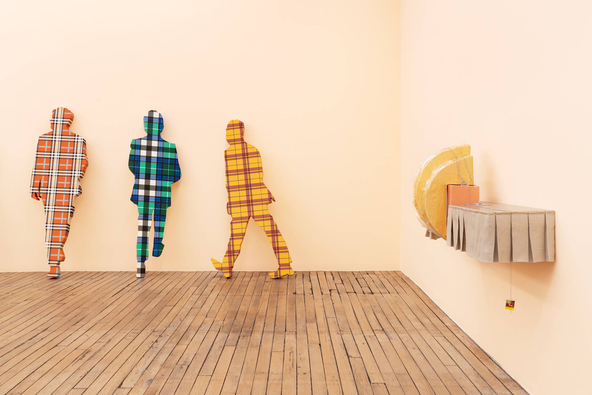 Five flat, life-sized human figures covered in plaid patterns lean against a peach wall in an art gallery. On the right wall, a shelf lined with a short, beige, pleated curtain holds two yellow cushions and an orange shoe box.