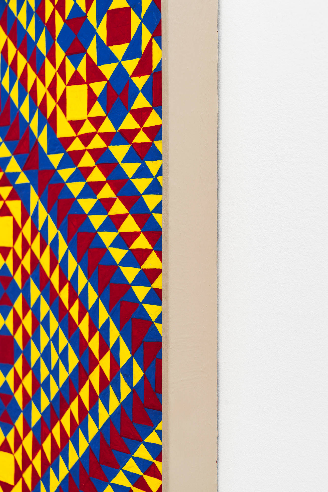 A detail shot of a colorful, symmetrical, geometric painting on board consisting of a pattern of repeating diamonds and triangles of yellow, red, and blue, in a tan artist frame.