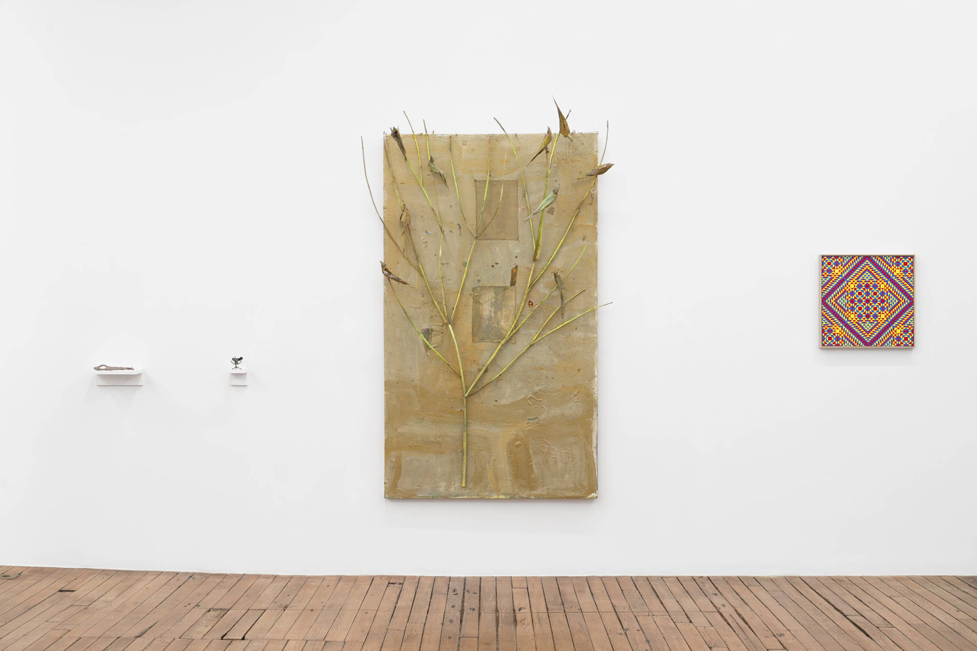 On the south wall of the gallery, a large gold painting by Tom Hoyt featuring a tree and birds sits on a white wall above a wood floor. To the right, there is a colorful, geometric painting by Matt Hoyt. To the left, 2 small sculptures by Matt Hoyt sit on white shelves.