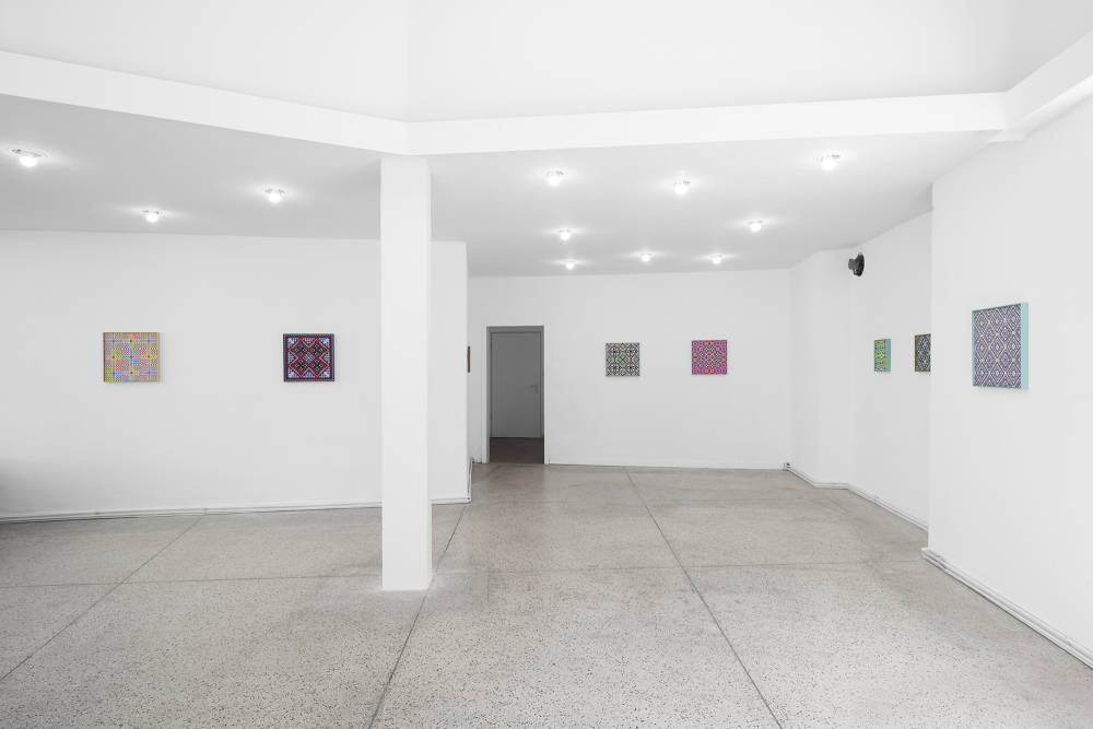 Image of a large gallery space that is well lit. There are eight colorful grid paintings installed on five walls.