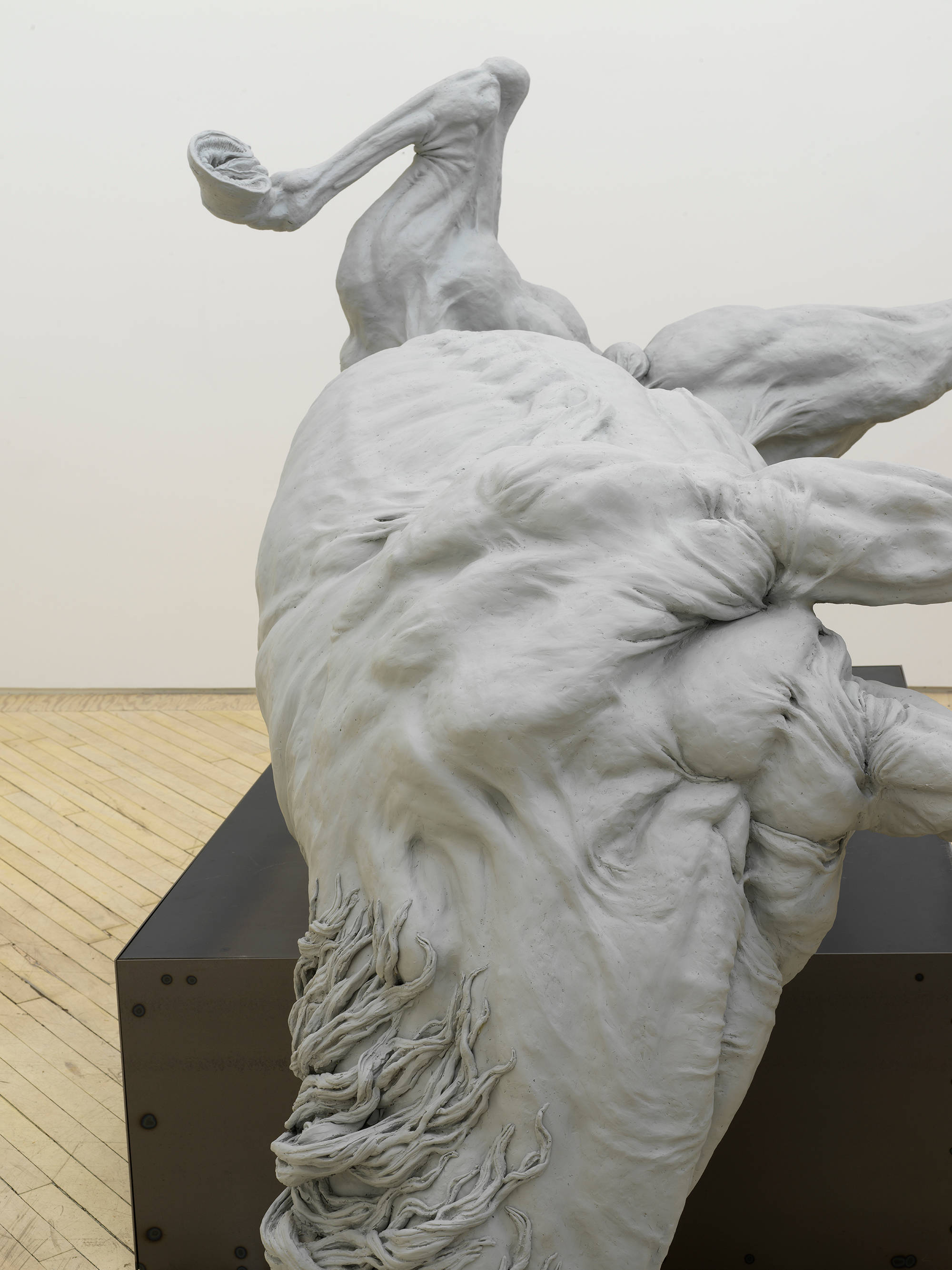 In a gallery space, a large sculpture of a fallen horse resting on top of an iron base.