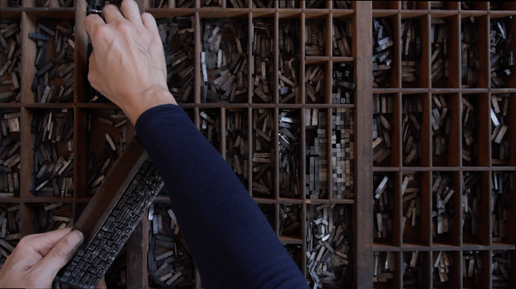 Still image from Ellie Ga's video 'Four Thousand Blocks' showing a close up of a wooden case for the lead block letters for typesetting print with the artists hand putting together a phrase from lead type to be set.