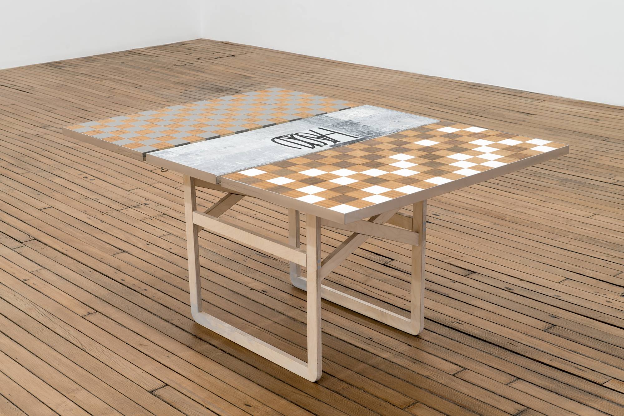 Ian Miyamura plywood table sculpture featuring a checkerboard top and removable leaf painting of depicting the letters "T, A, S, T, E" arranged at different sizes and angles on a light grey background.