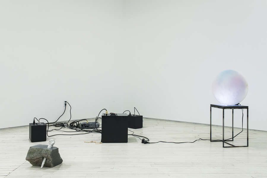 In the corner of a gallery there are a series of three speakers directly on the floor, wires messily strewn about, to the left is a sculpture of a large chunk of stone with a cast metal microphone piercing it, to the right a sculpture of a metal and stone plinth with a cylindrical piece of alabaster, lit from behind by a lightbulb, with airbrushed tones of pink and blue on the face. 