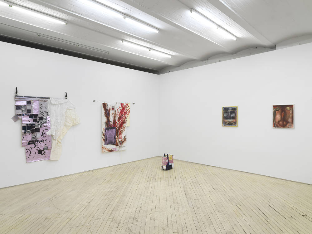 In a gallery space, to the left are two irregularly shaped abstract paintings. On the floor in the corner a small sculpture resembling a boat. To the right are two small paintings in metal frames. 