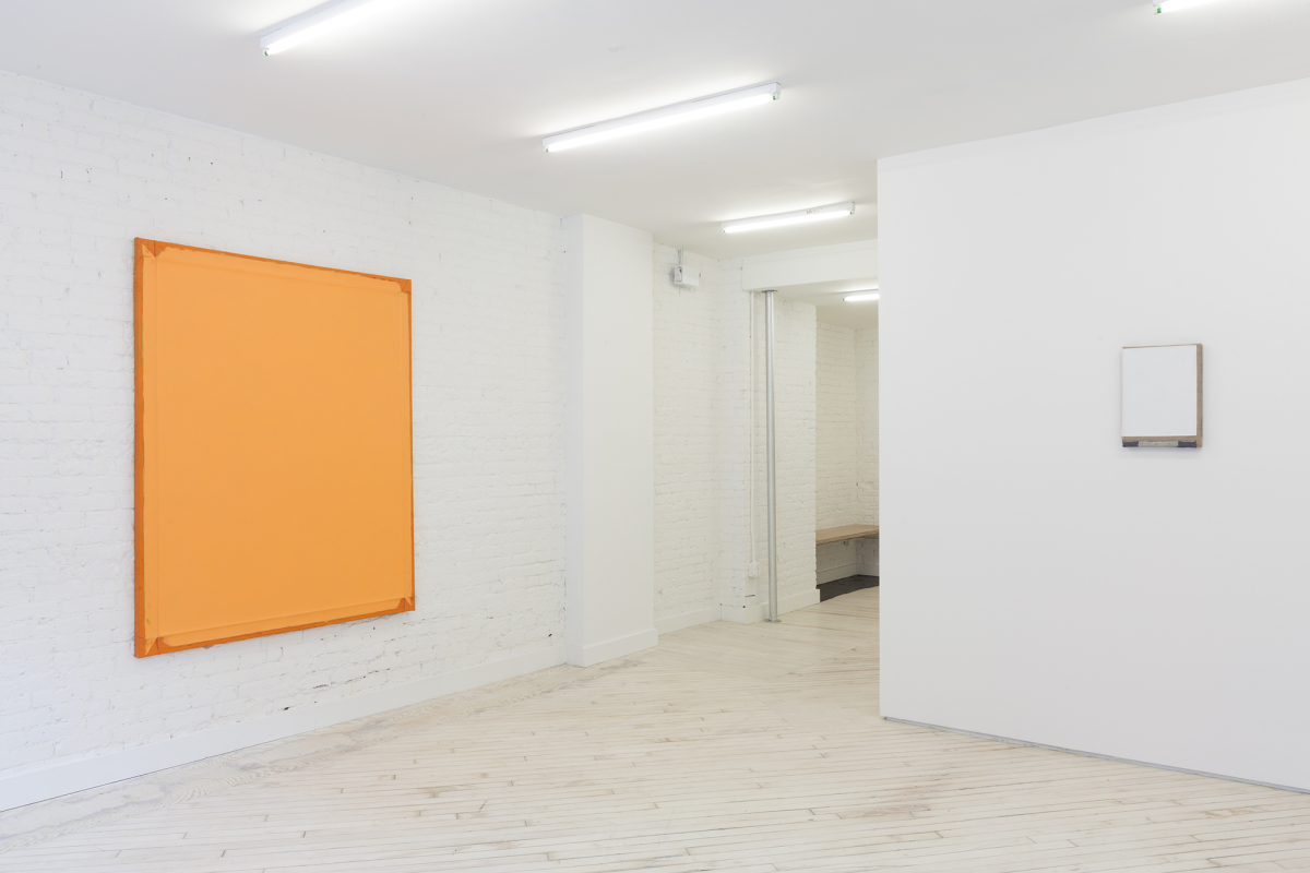 View of front lobby gallery at Bureau - at left a large orange monochrome painting hangs on the brick wall, at right, a small white painting with grey stripes at the bottoms hangs on the wall.