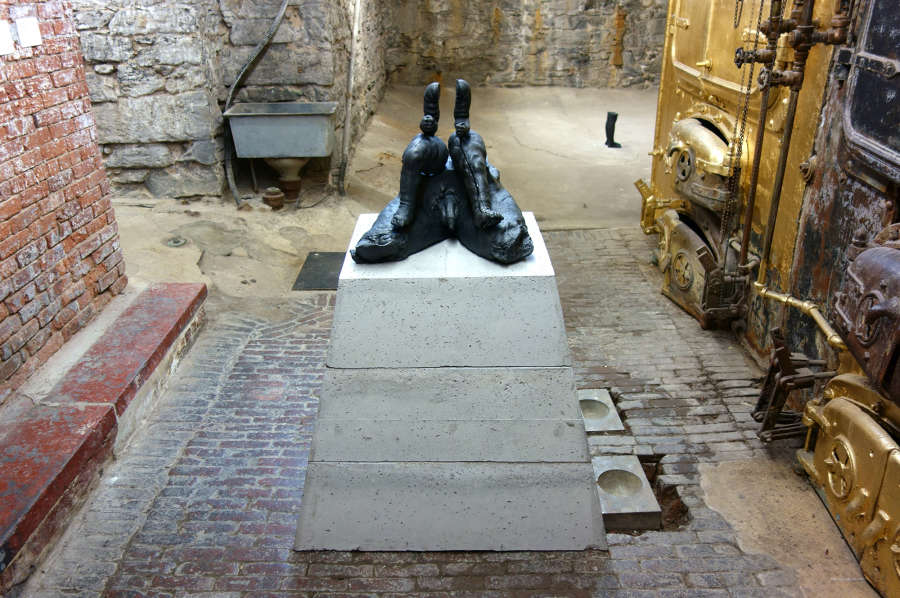 Installation view of metal and concrete sculpture with a trapezoidal base, containing a cast iron sculpture on top depicting a man and child's legs and disembodied child's arms. 