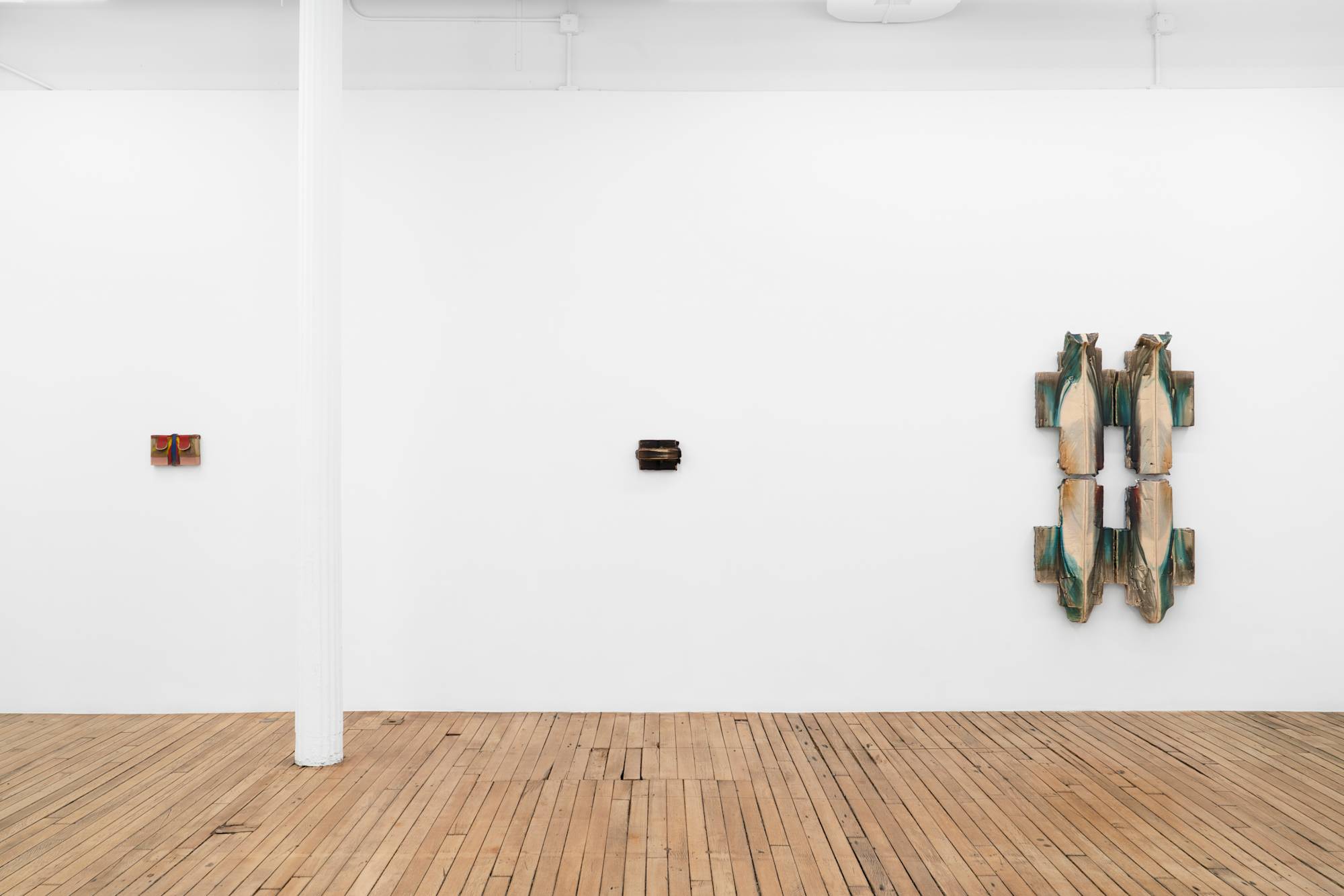 Three wall-mounted ceramic sculptures of various color and scale hang in a line on a white wall in an art gallery with a white column and wooden floor.