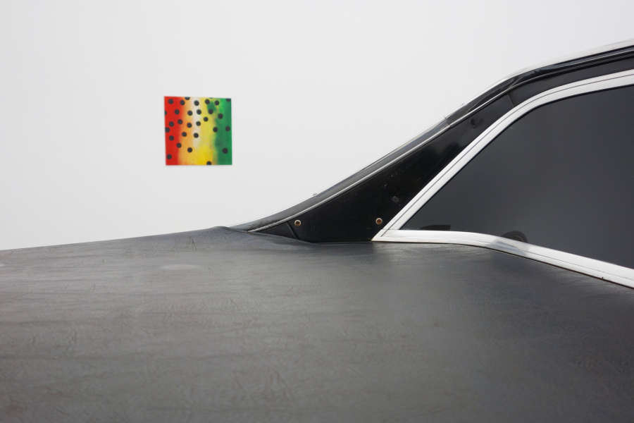 A cropped image of a black vintage car in the foreground. In the background is a small abstract painting with red, yellow, and green stripes of color. On the surface are numerous painted black dots. 