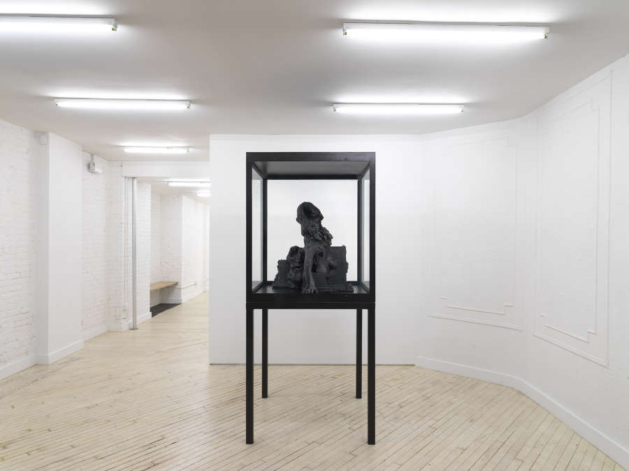 Installation view of metal and glass vitrine on stilts containing a sculpture made of cast iron depicting a decaying figure.
