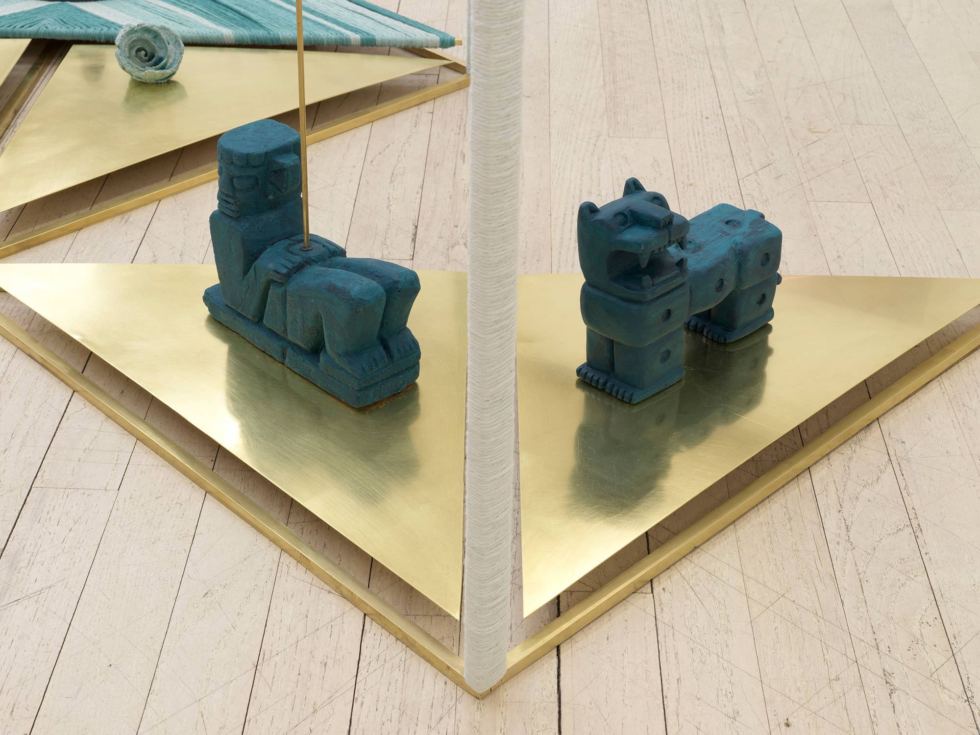 Image of a sculpture by Claudia Peña Salinas composed of vertical brass rods creating rectangular shapes in space with parts of the rod wrapped in dyed blue fabric. Sitting on the base of these rectangles are small sculptures of Mayan symbols.