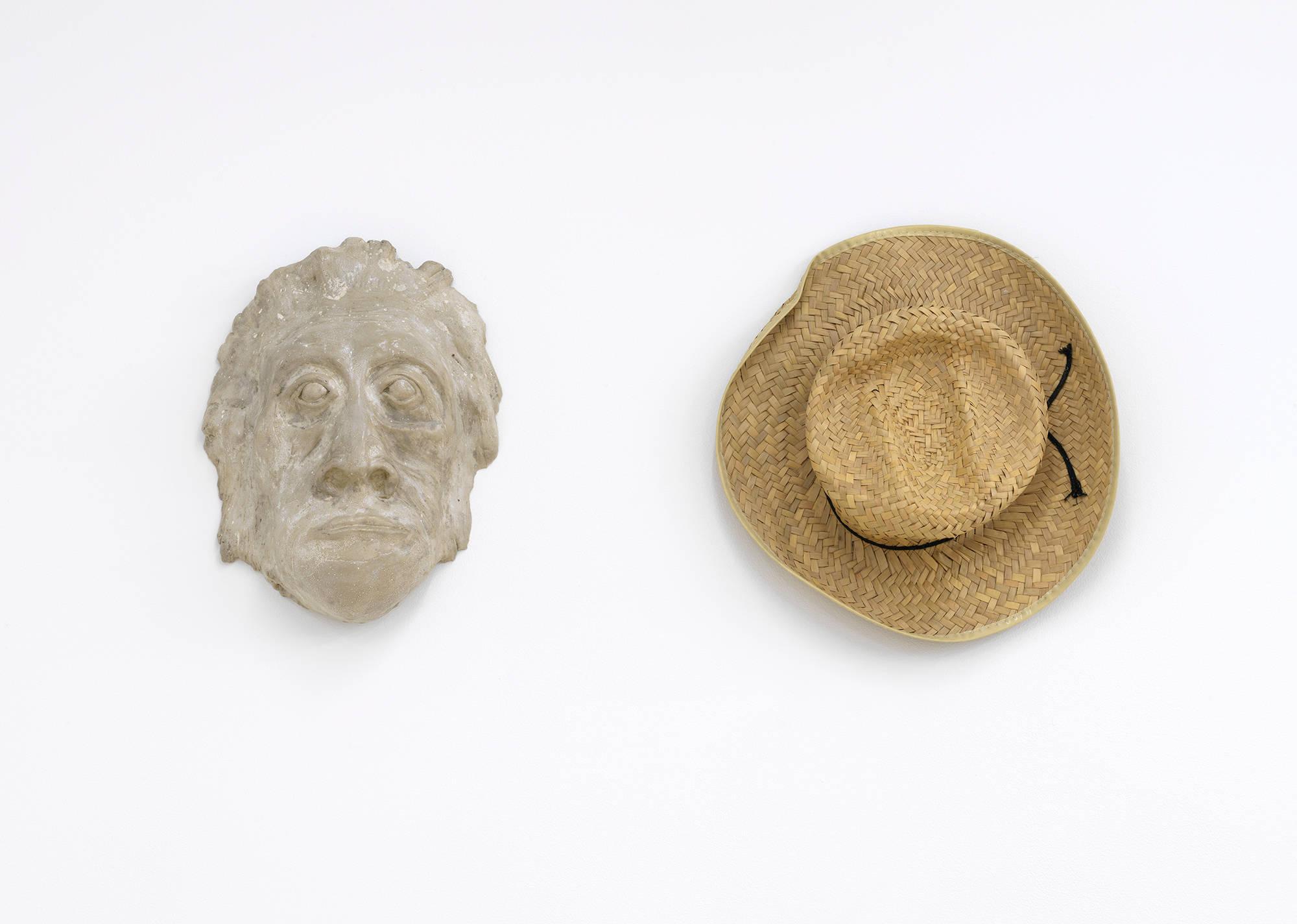Image of a mask made of clay installed on the left, next to a straw brim hat, hanging on the wall.