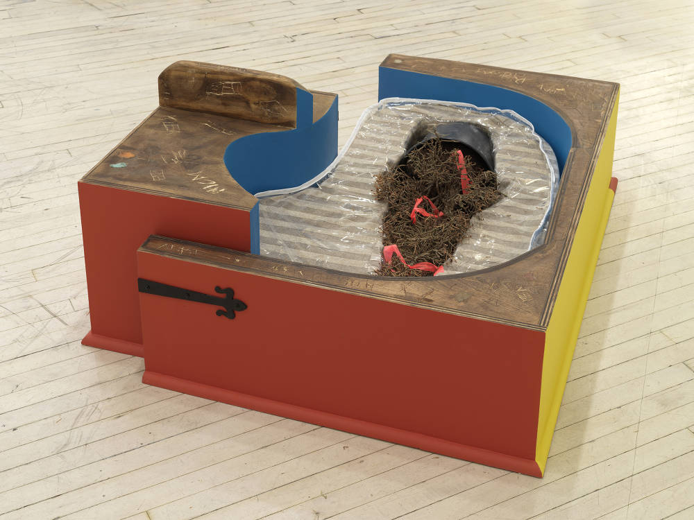 Image of an artwork by Libby Rothfeld of two conjoined boxes, one taller than the other, with red blue and yellow paint on the exterior of the box. There is a dead Christmas tree in a circular cut out of the center of the box. The Christmas tree is embedded into what looks like a couch cushion with a plastic cover. There is graffiti scratched into the surface of the wooden box and bubblegum applied to it.