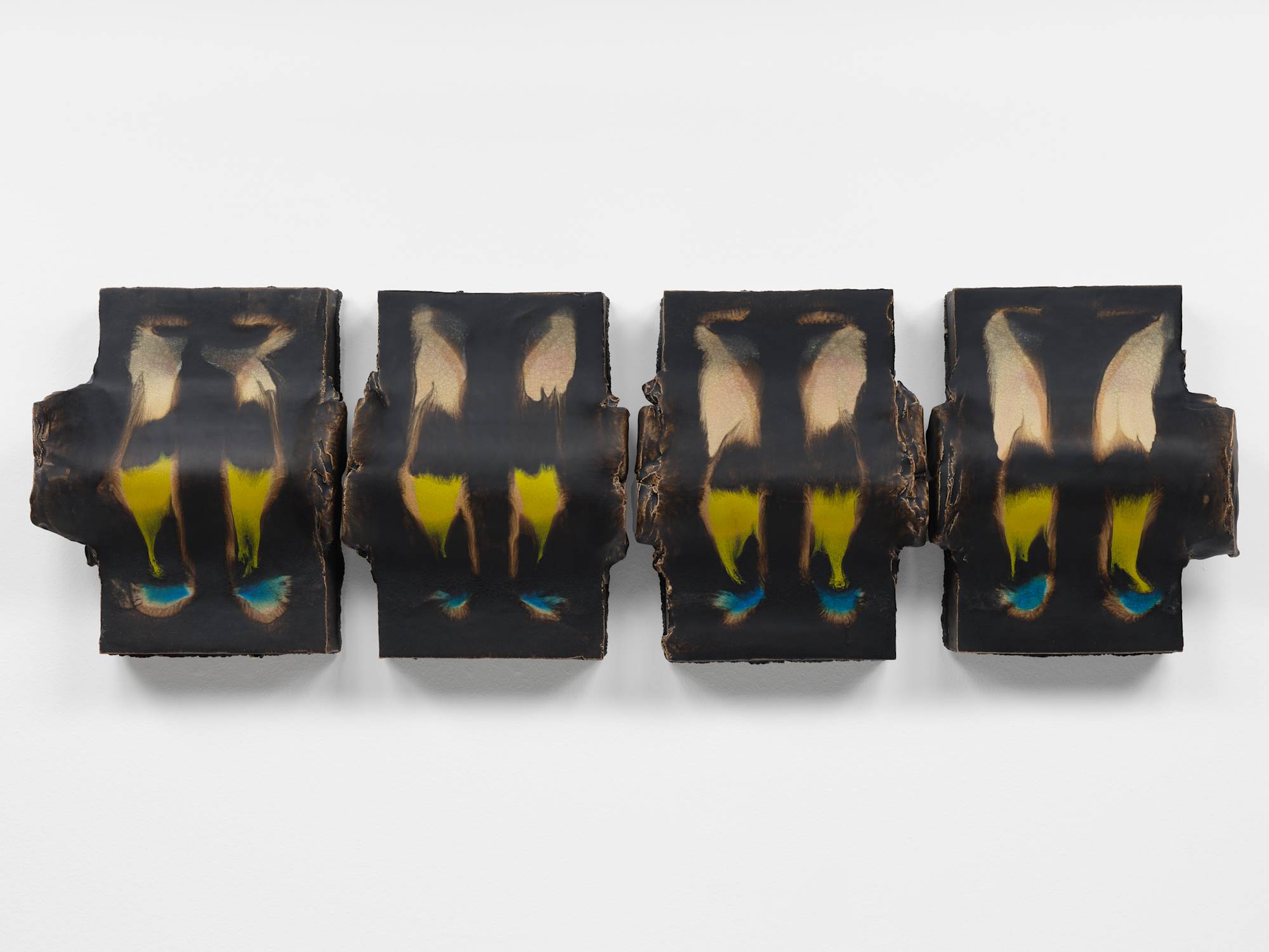 Four nearly identical vertical cuboid ceramic sculptures hang in a tight horizontal line on a white wall. Each piece has two horizontal cylindrical bands dividing their composition beneath the smooth top surface that has been glazed dark brown with accents of tan, green, and blue.