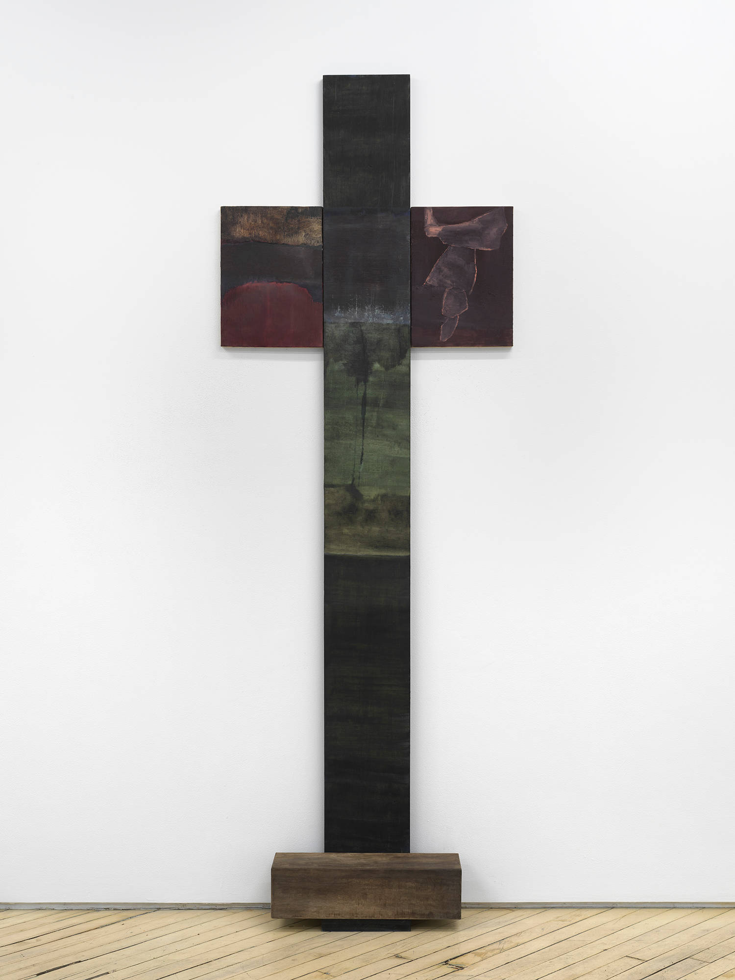 A vertical painting and sculpture in the shape of a cross leaning against a gallery wall.
